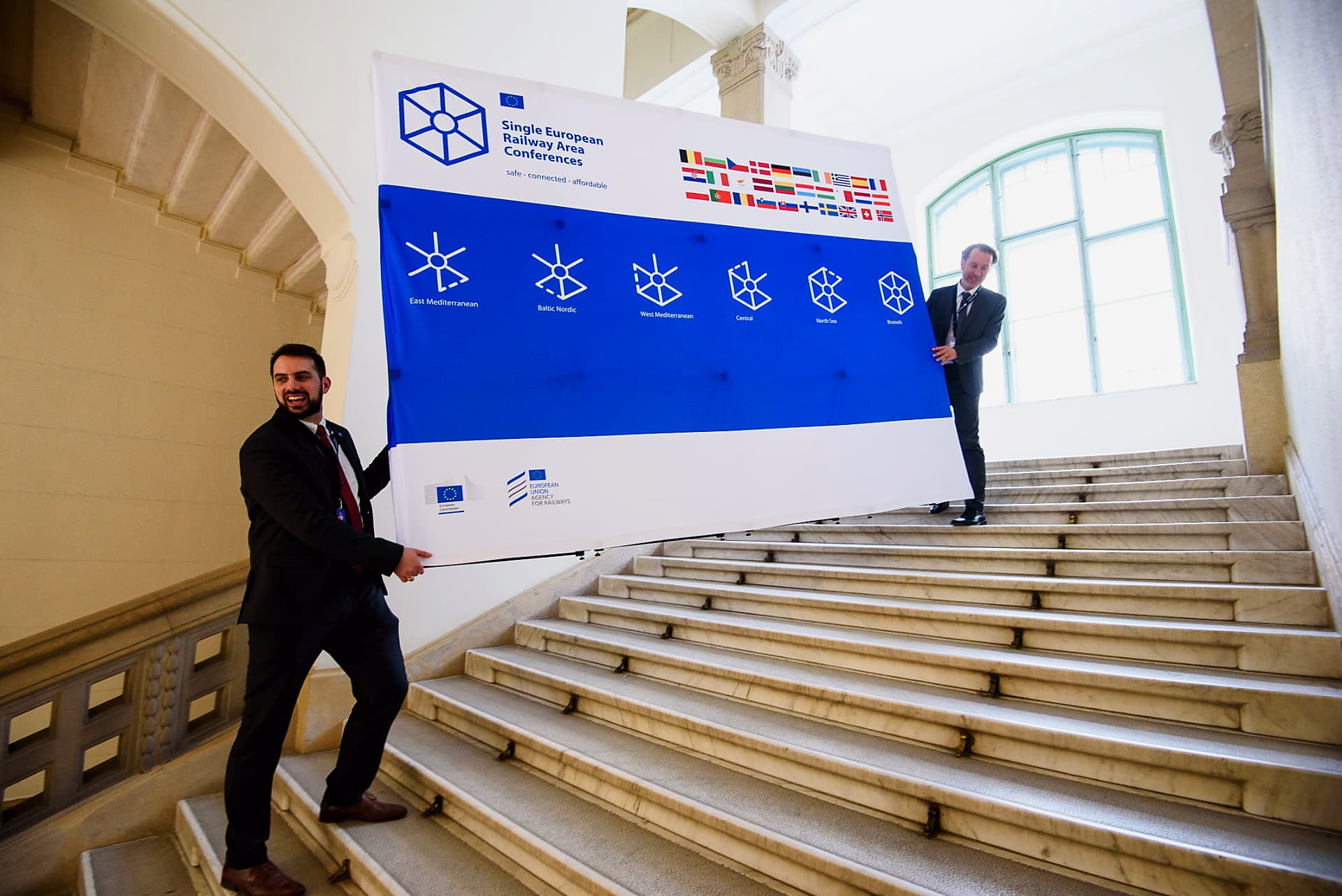Two European Union Agency for Railways staff carrying the large Single European Railway Area Conferences banner up a marble staircase in Brussels