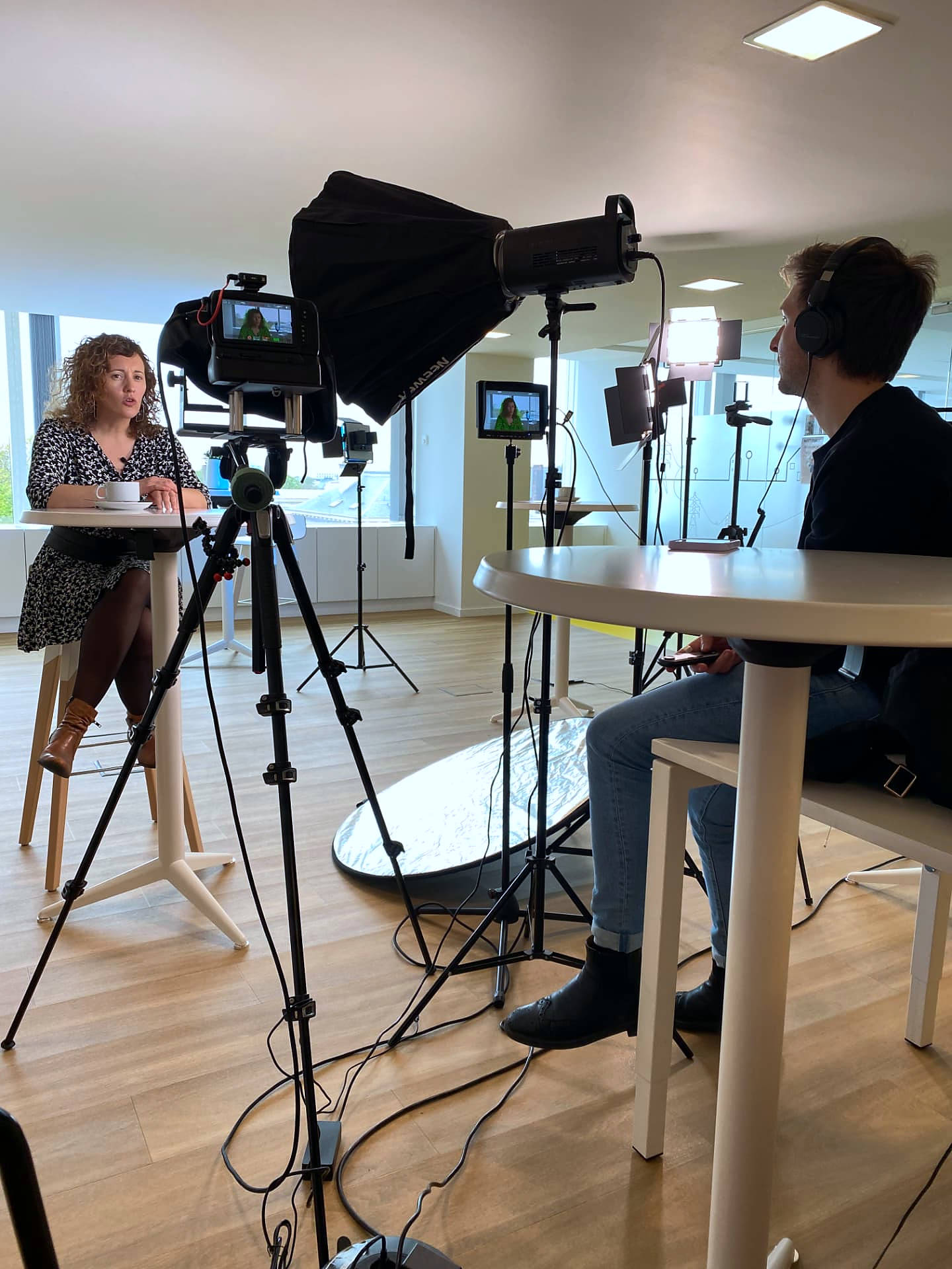 A recorded conversation in progress: two people seated at high tables with camera, monitor and lighting between them