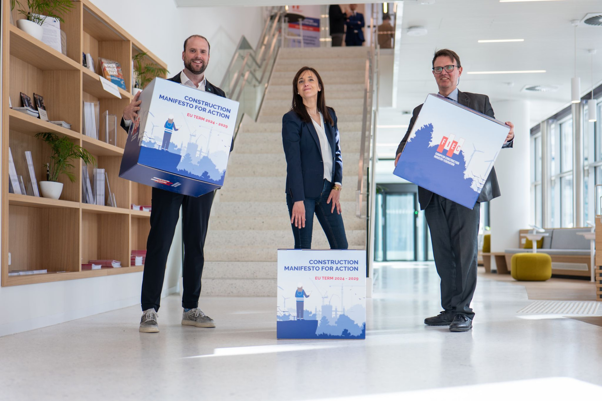 Three representatives hold large cubes forming the FIEC Construction Manifesto for Action — EU Term 2024-2029 campaign produced by #inextremis, in a bright Brussels atrium