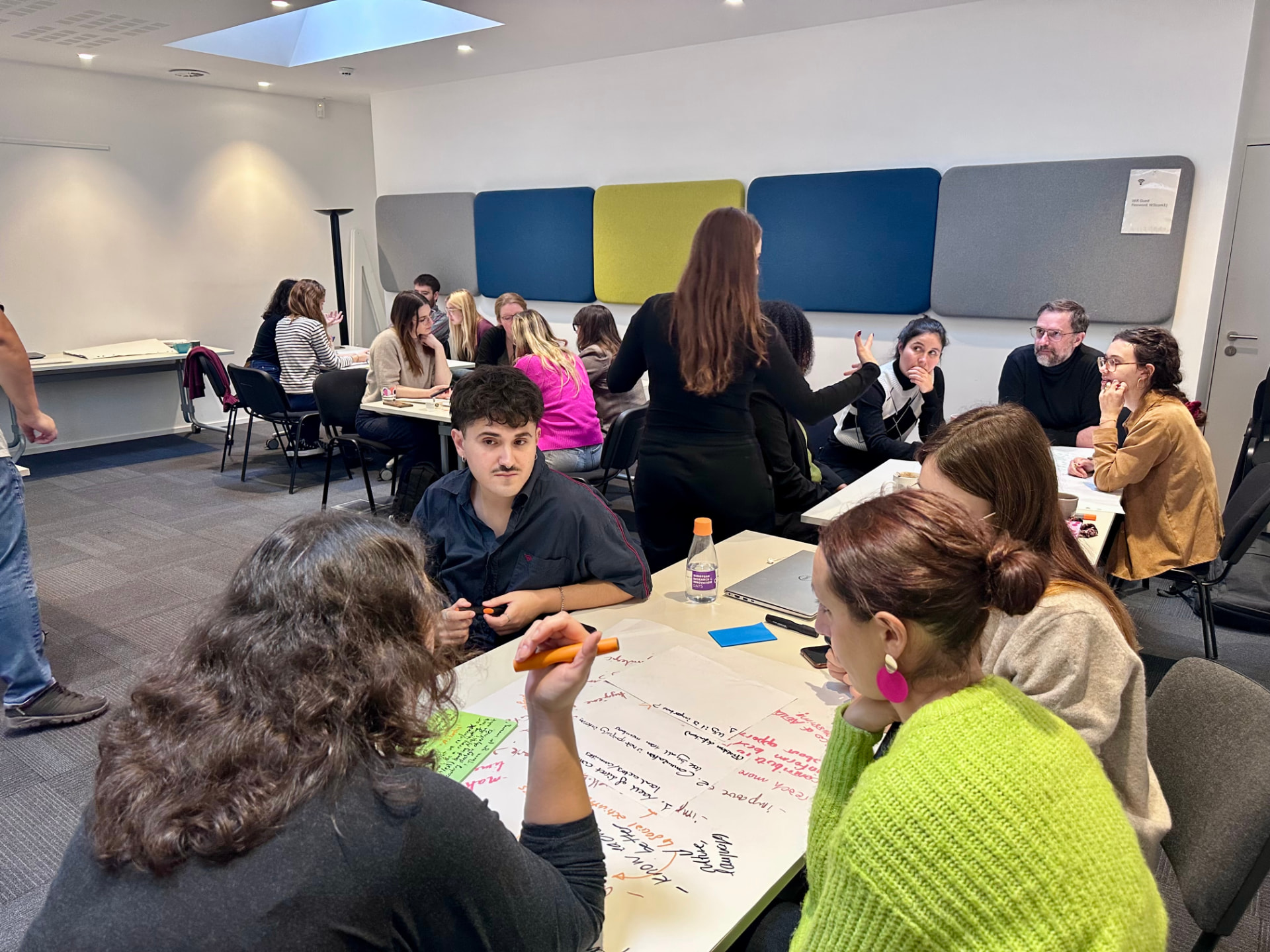 A facilitated #inextremis workshop in progress: participants at several tables annotating large sheets with priorities and actions while a facilitator leads the room