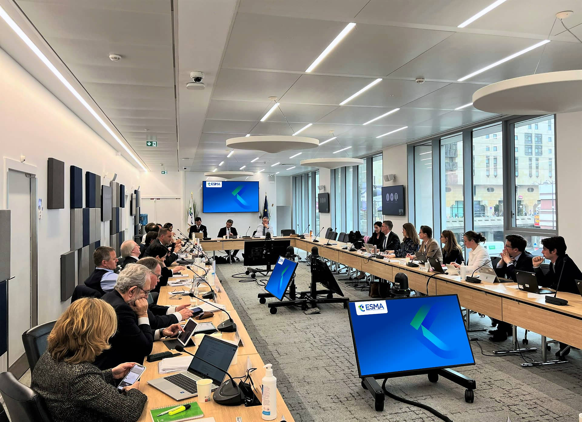 An ESMA stakeholder meeting in a bright Brussels conference room: participants seated around a U-shaped table with ESMA-branded screens and the new identity visible