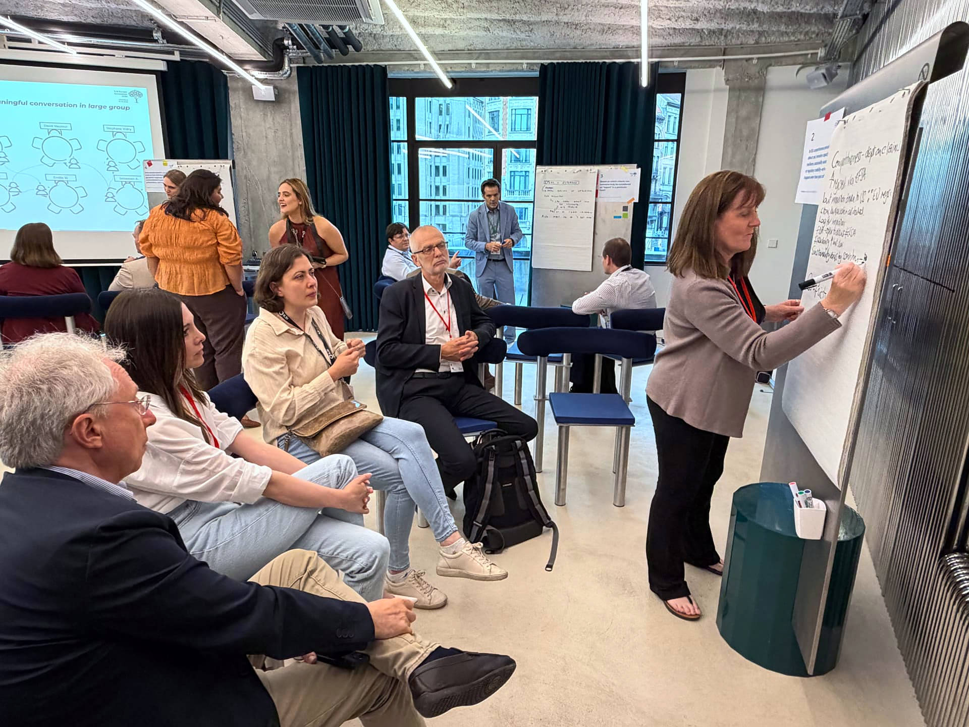 A facilitated strategy workshop in progress: a participant captures group input on a whiteboard while others follow a large-group conversation framework projected behind her